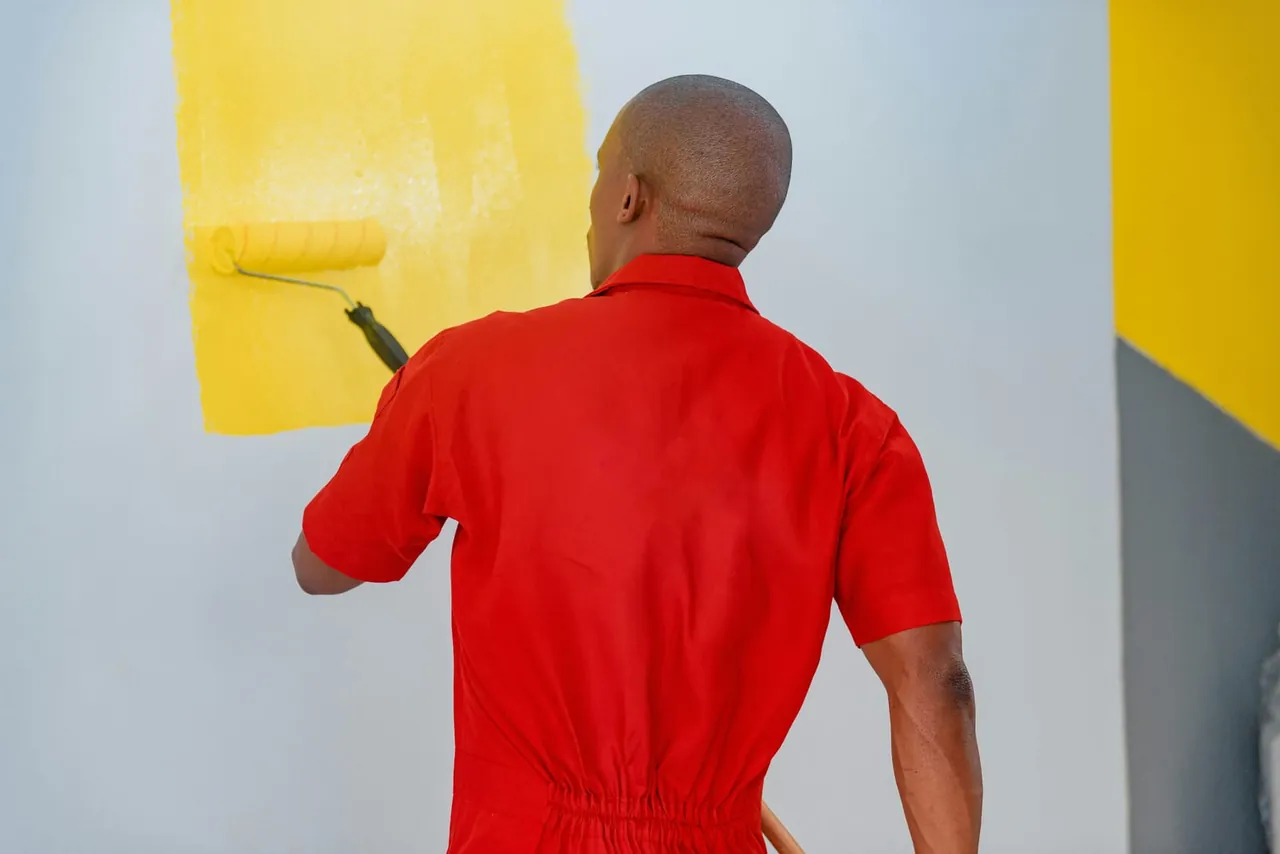 Malerfachkraft in roter Arbeitskleidung beim Anstrich einer Wand mit gelber Farbe